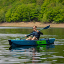 Man paddling a blue kayak on a calm lake with green trees in the background