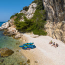 People sitting on a rocky beach with kayaks nearby, surrounded by cliffs and clear blue water.