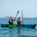 Two people kayaking in a blue kayak on calm water with mountains in the background.